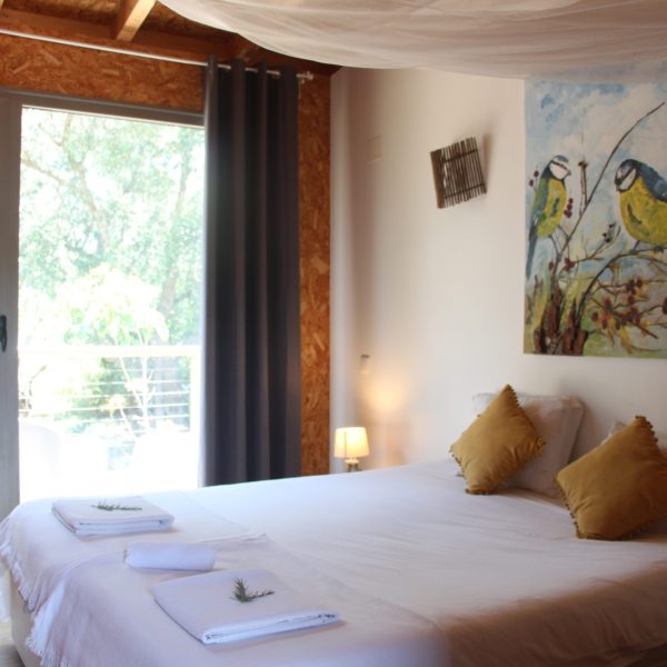 Quarto luminoso com cama de casal, almofadas mostarda e decoração natural, incluindo uma pintura de aves e ramos de alecrim sobre as toalhas. A janela ampla oferece vista para o exterior arborizado.