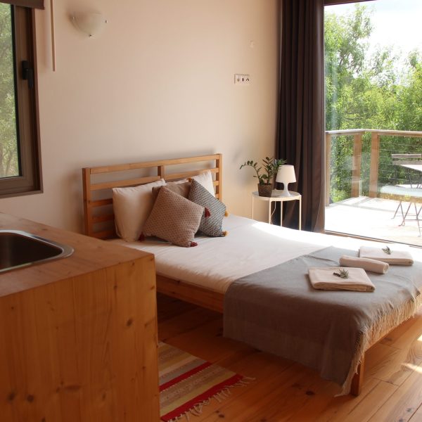 Quarto com kitchenette integrada em madeira, cama de madeira com roupa de cama branca e manta azul-clara, duas almofadas decorativas, e tapete colorido no chão. Ambiente luminoso, com janelas grandes e elementos naturais.