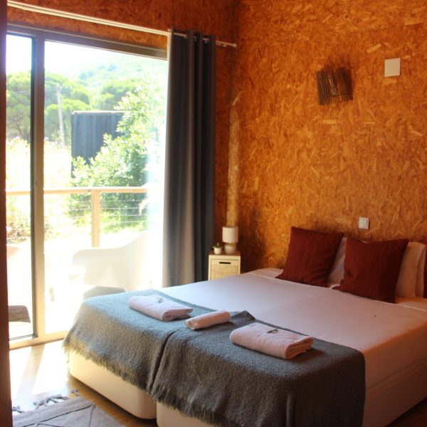 Quarto com cama de casal coberta por manta cinzenta e almofadas castanhas, junto a uma parede de madeira OSB. À esquerda, portas de vidro abertas revelam uma varanda com cadeiras brancas e vegetação exterior iluminada pela luz natural.