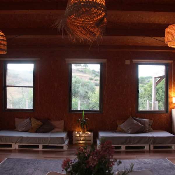 Sala ampla e acolhedora com janelas voltadas para o verde exterior, sofás de pallet com almofadas e iluminação suave em um espaço de descanso ecológico.