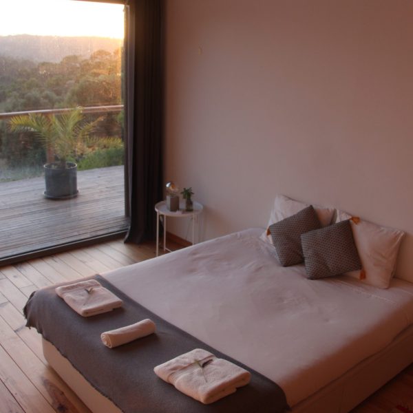 Quarto acolhedor com teto em madeira exposta, cama feita com roupa branca, mesa de apoio com candeeiro e janela panorâmica com vista para a natureza.