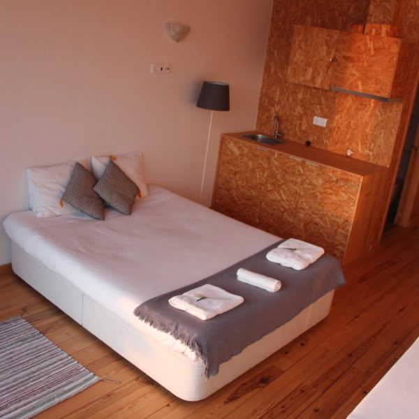 Espaço integrado com cama confortável e pequena cozinha, ideal para estadias práticas num ambiente acolhedor com acabamentos em madeira.