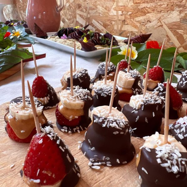 Close de docinhos de banana, morango e chocolate com coco, dispostos em tábua rústica, acompanhados por jarra de smoothie natural e pratos coloridos ao fundo.