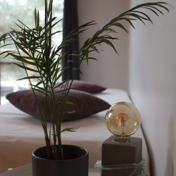 Detalhe de uma mesa de apoio com uma planta em vaso preto e um candeeiro de base em cimento, junto a uma cama com almofadas em tons terra.