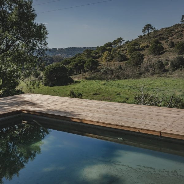 Deck de madeira junto a uma piscina com água cristalina, oferecendo vista para um vale verdejante e montanhoso, ideal para relaxar em harmonia com o ambiente natural.