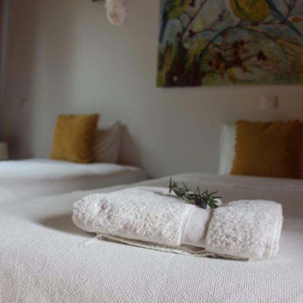 Quarto com duas camas individuais cobertas por mantas brancas e almofadas mostarda. Um ramo de alecrim decora uma toalha cuidadosamente dobrada sobre a cama em primeiro plano.
