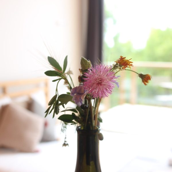 Vaso de vidro com flores silvestres em destaque, ao centro da imagem, com o fundo desfocado revelando uma cama com almofadas decorativas e uma janela com cortina aberta, permitindo a entrada de luz natural.