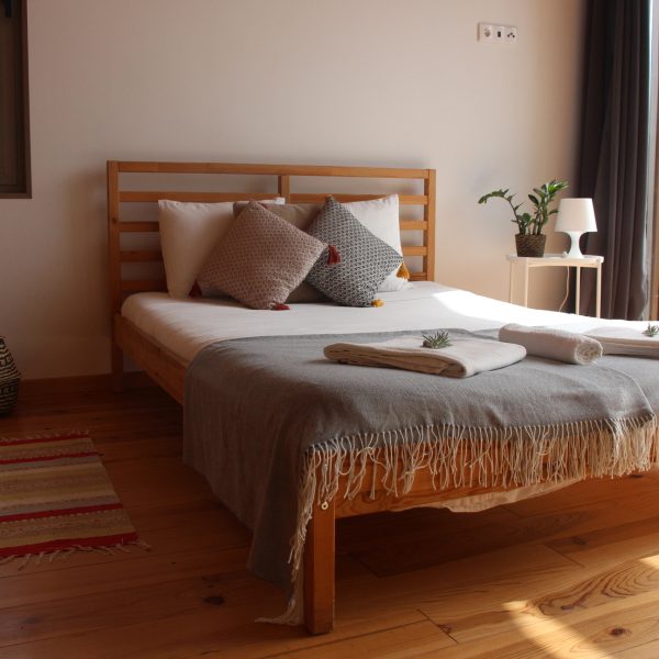 Vista ampla de um quarto acolhedor com cama de madeira coberta com manta cinza-clara e almofadas decorativas. A luz natural entra por uma janela ampla com vista para a vegetação exterior. Ao lado da cama há uma mesa branca com candeeiro e planta decorativa.