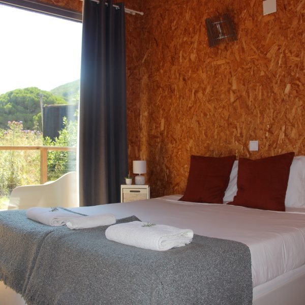 Quarto acolhedor com decoração em tons naturais, onde uma cama bem preparada convida ao descanso. A luz suave entra pelas portas envidraçadas, revelando uma varanda com vista para o jardim e as colinas verdes ao fundo.