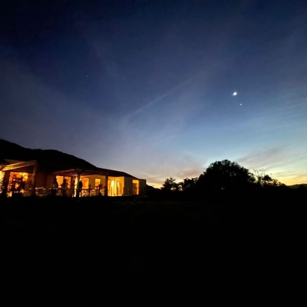 Fachada acolhedora casa ecológica iluminada ao anoitecer, rodeada por natureza, sob um céu azul profundo com estrelas e a lua ao fundo.