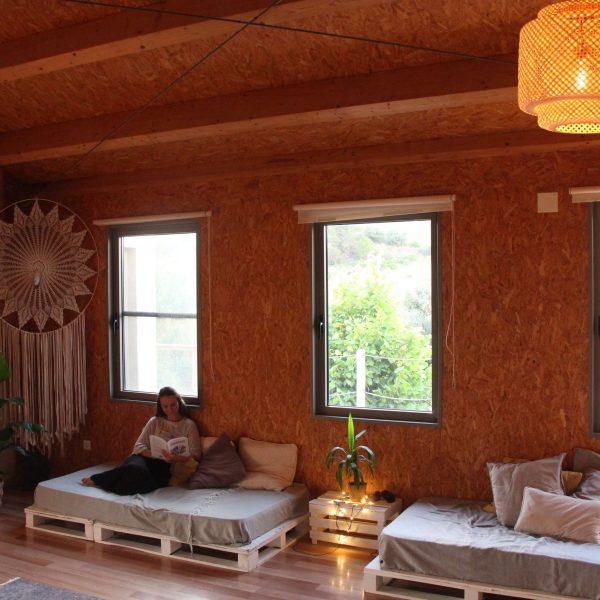 Espaço interior tranquilo com uma pessoa relaxando e lendo, rodeada por decoração artesanal e natureza visível pelas janelas.
