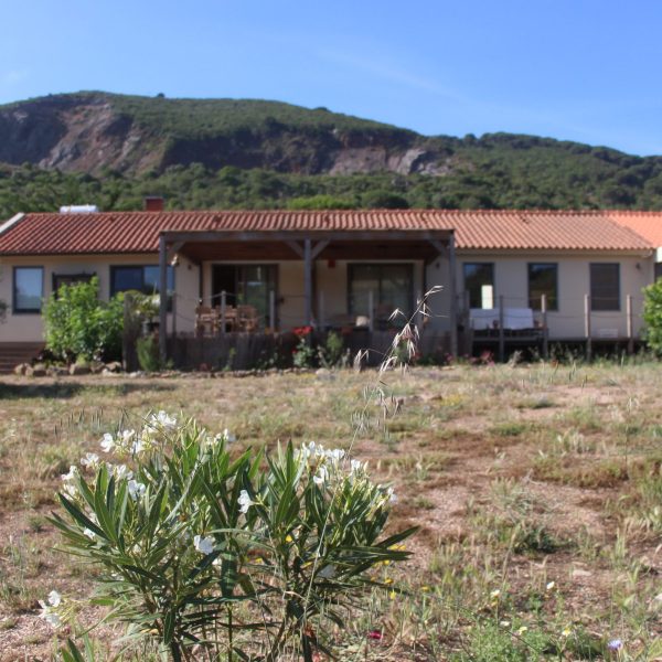 Casa ecológica com fachada clara, telhado tradicional e jardim florido, com vista para colinas verdes e paisagem montanhosa.