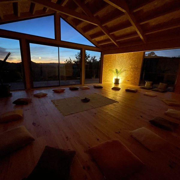 Sala de madeira com janelas panorâmicas e almofadas dispostas em círculo, ideal para meditação ou encontros ao entardecer com vista para as colinas.