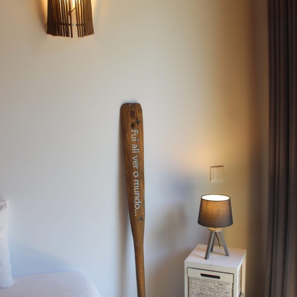 Pormenor decorativo de um quarto: iluminação de parede rústica, mesa de cabeceira com candeeiro moderno e remo em madeira com inscrição inspiradora.