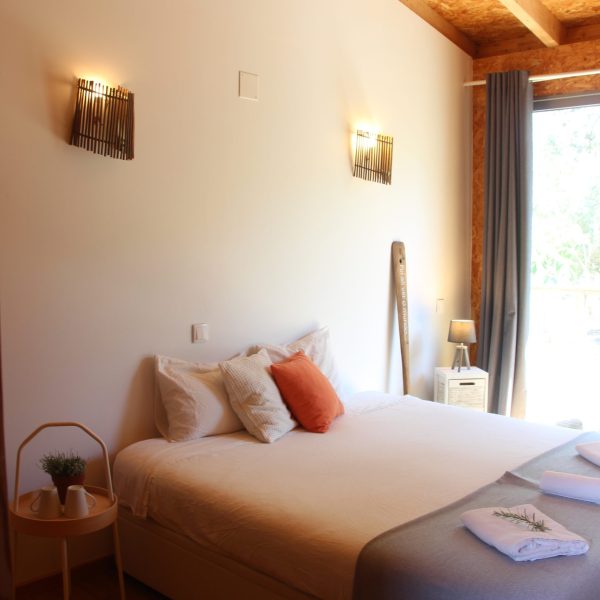 Vista ampla de quarto com design natural e acolhedor, iluminação suave, cama com almofadas coloridas e varanda rodeada por natureza.