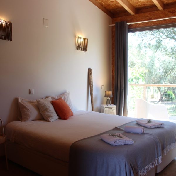 Quarto acolhedor com cama de casal, almofadas terracota e manta cinzenta, junto a uma janela ampla com vista para a vegetação exterior.