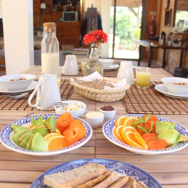 Pequeno-almoço saudável servido sobre uma mesa de madeira com pratos de fruta fresca colorida, pão caseiro, compotas e leite, num ambiente acolhedor com luz natural.